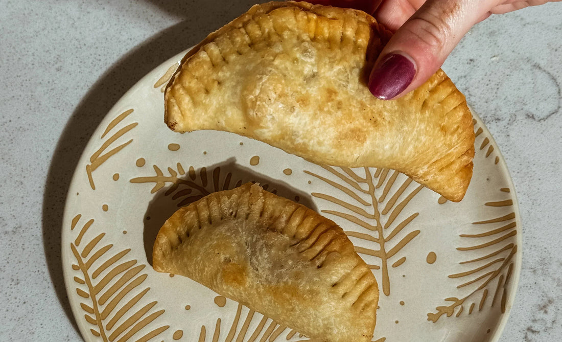 Hand holding a golden-brown empanada on decorative plate, showcasing Mora's artisanal food.