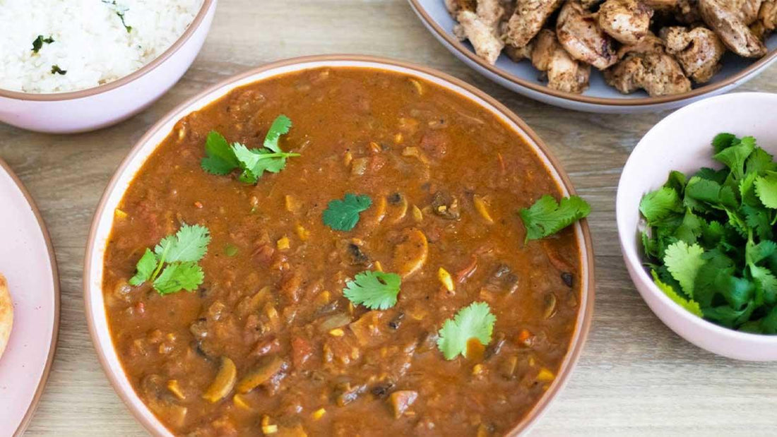 Bowl of vegetable curry garnished with cilantro, served with rice, grilled chicken, and fresh cilantro, on a wooden table