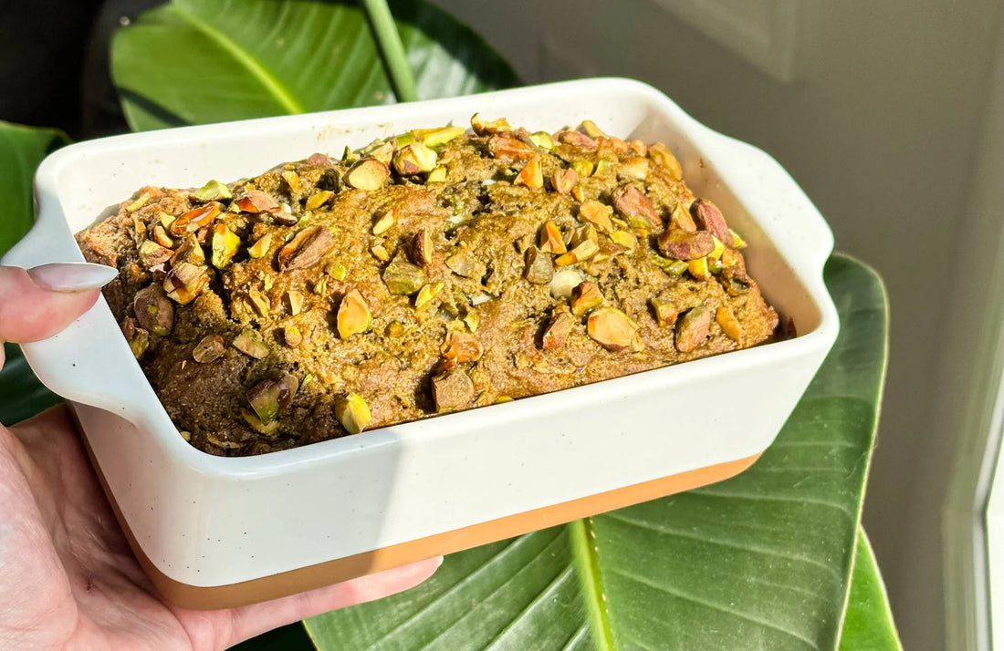Freshly baked pistachio loaf in a ceramic dish, held above green leaves in natural light