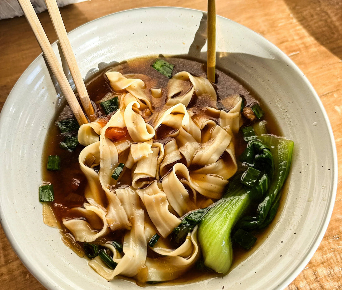 Bowl of noodle soup with bok choy, scallions, broth, and wooden chopsticks on table